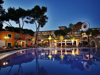 Osvětlený bazén v noci v hotelu obklopeném stromy a barevnými budovami.