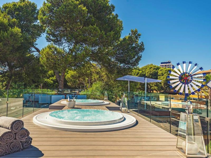 Dřevěná terasa s vířivkou, ručníky, slunečníkem, bazénem a stromy pod modrou oblohou.