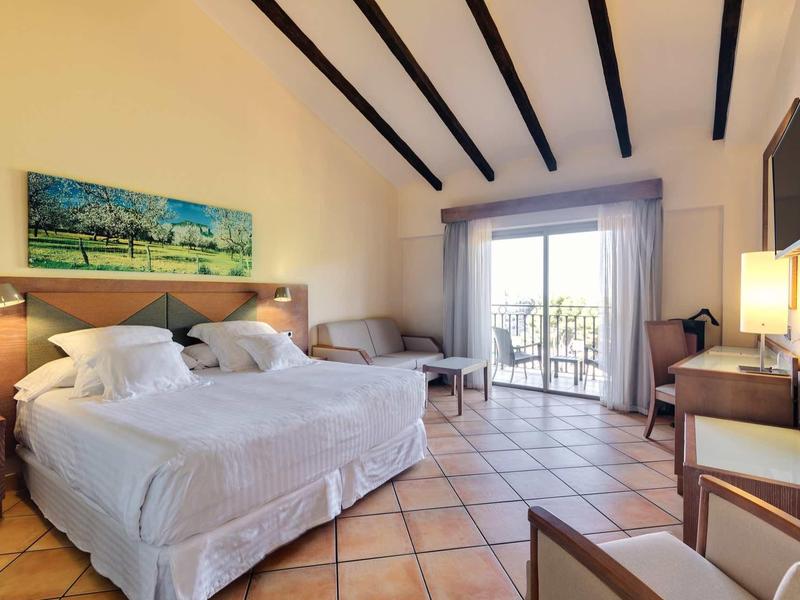 Prostorný hotelový pokoj s velkou postelí, křesly, balkonem a výhledem ven.