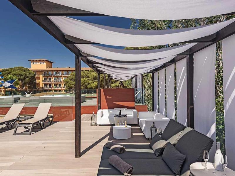 Hotelová terasa s lehátky a pohovkami pod bílou pergolou u bazénu.