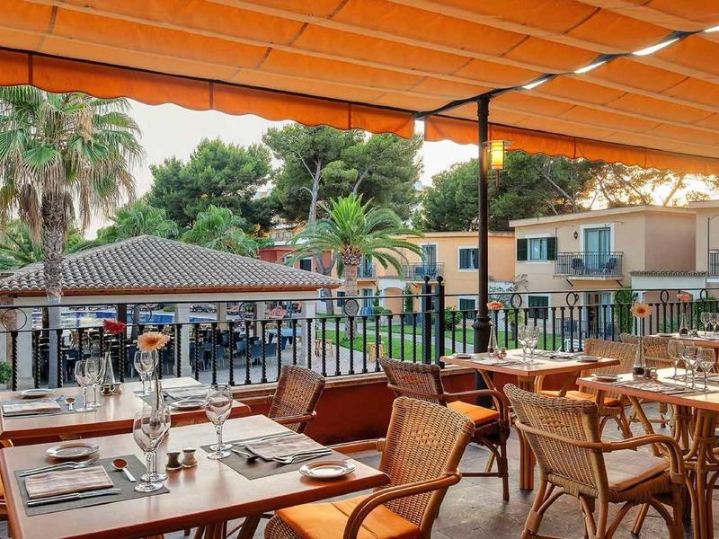 Restauranční terasa s výhledem na bazén obklopený palmami pod oranžovou pergolou.