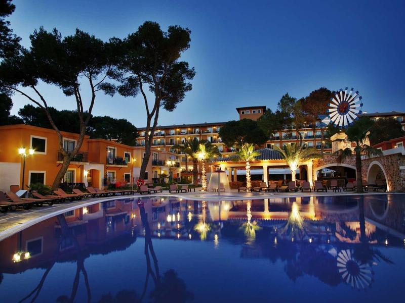 Osvětlený bazén v noci v hotelu obklopeném stromy a barevnými budovami.