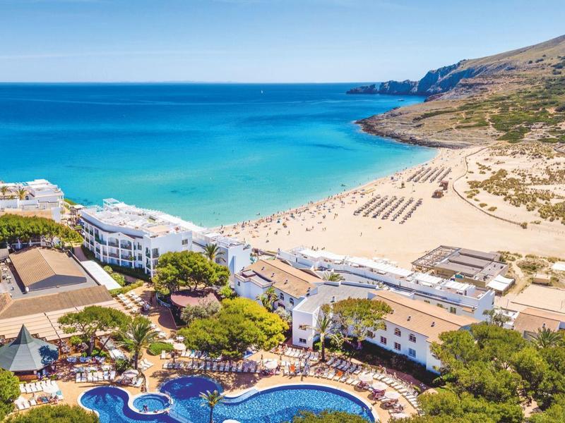 Vista di un resort hotel con piscina e spiaggia sabbiosa lungo una costa collinare.