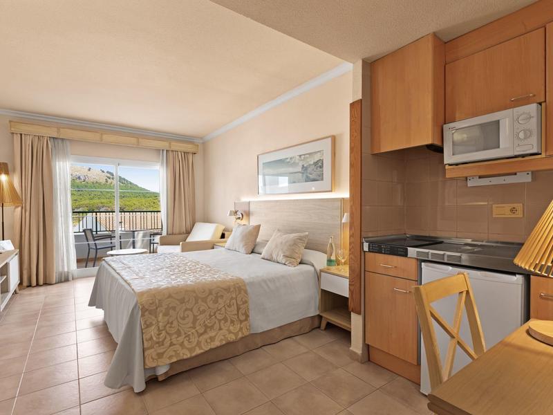 Camera d'hotel luminosa con letto matrimoniale, angolo cottura e balcone con vista montagna.