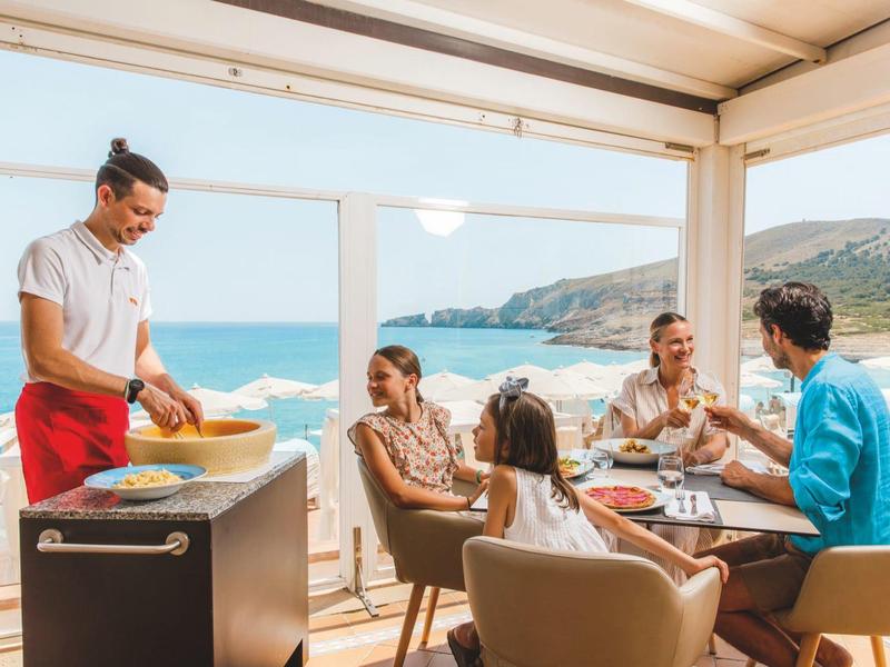 Famiglia seduta al tavolo del ristorante con vista sul mare, cameriere che serve il cibo.