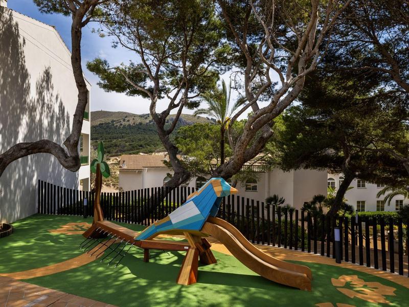 Area giochi per bambini con scivolo e struttura per arrampicata sotto gli alberi accanto a un edificio bianco.