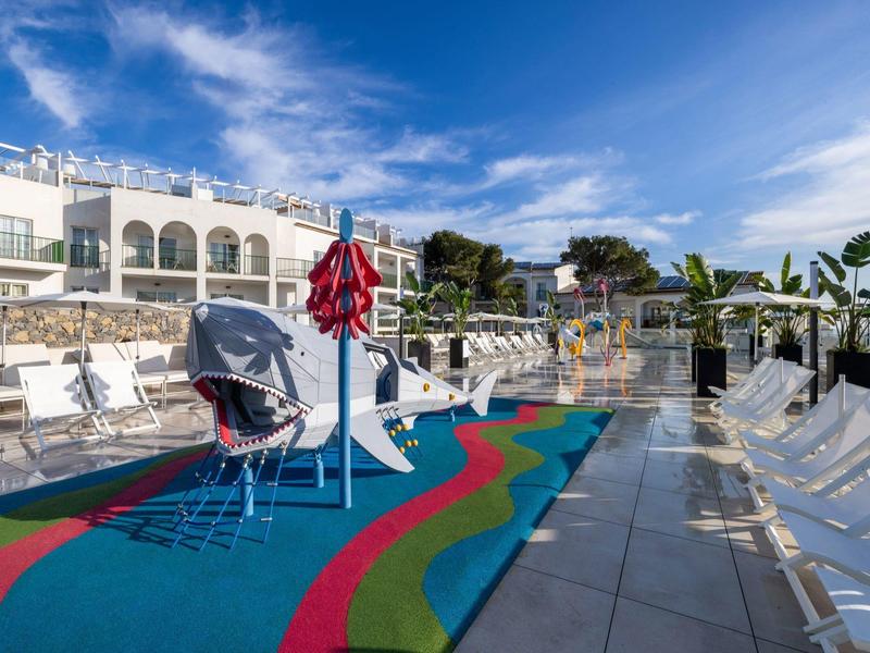 Un'area piscina esterna luminosa con sedie a sdraio, pavimento colorato e ombrelloni sotto un cielo azzurro.