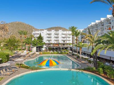 Hotelanlage mit Pools, Liegestühlen und Palmen vor bergiger Kulisse unter blauem Himmel.