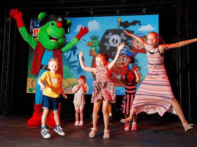 Kinder springen fröhlich auf einer Bühne mit einer grünen Maskottchen-Figur im Hintergrund.
