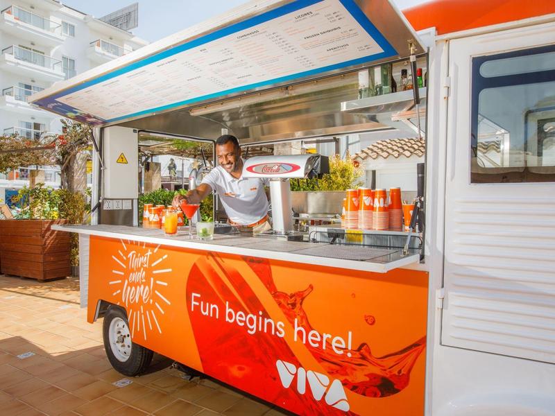 Foodtruck mit offener Theke und Mitarbeiterin, der frische Speisen serviert, im Freien bei Sonnenschein.