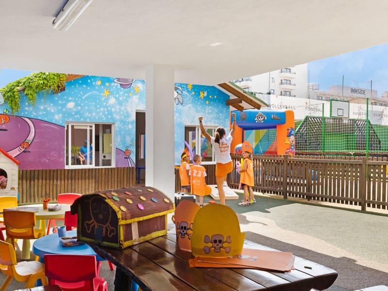 Ein überdachter Spielbereich mit bunten Stühlen, Tischen und Kinderhütten im Freien.