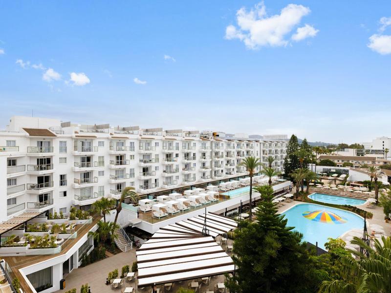 Großes Hotel mit weißem Gebäude, Swimmingpool und Sonnenliegen unter blauem Himmel.