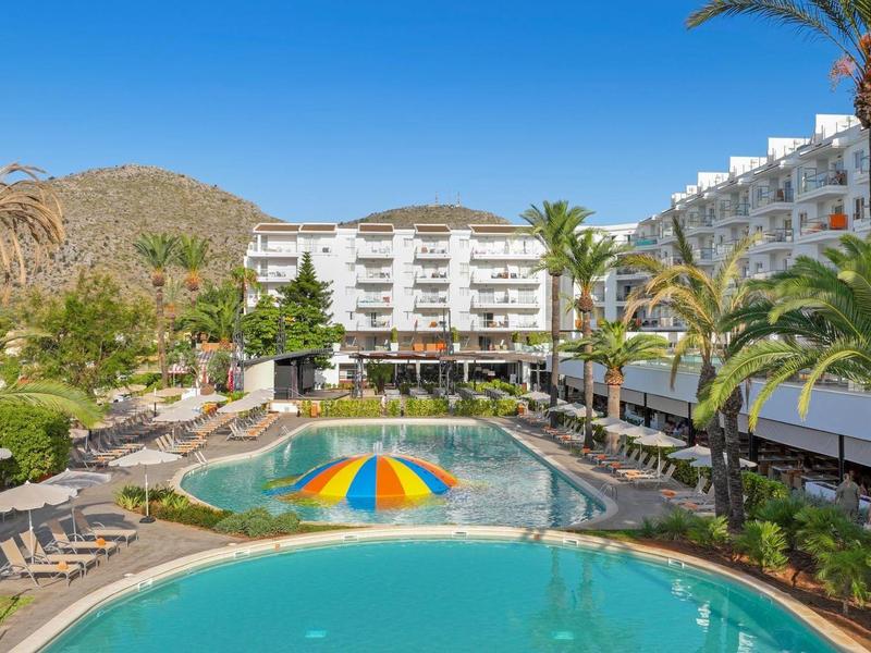 Hotelanlage mit Pools, Liegestühlen und Palmen vor bergiger Kulisse unter blauem Himmel.