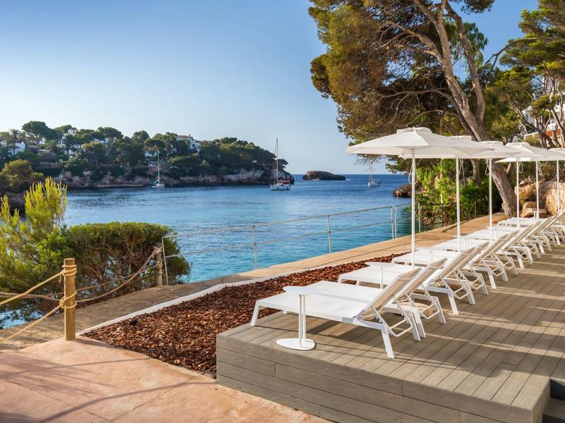 Bílé lehátka vedle sebe u zátoky s borovicemi pod jasnou oblohou.