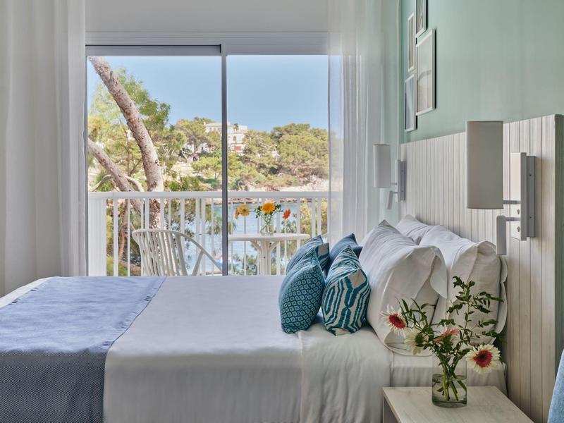 Světle hotelový pokoj s dvoulůžkem, balkonem a výhledem na zelené stromy.