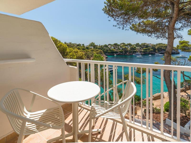 Balkon hotelu s kulatým stolem a židlemi, výhled na moře a zeleň.