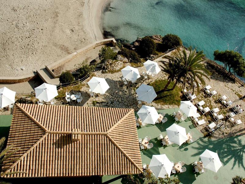 Letecký pohled na pláž s bílými slunečníky a palmami u budovy.