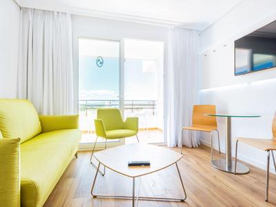 Salon lumineux avec canapé jaune, chaises, petite table et balcon avec vue sur la mer.