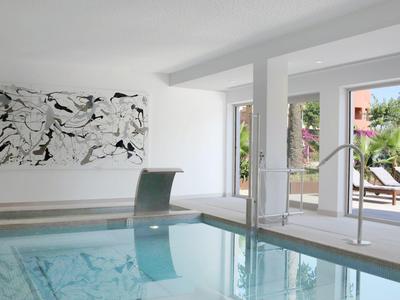 Piscine intérieure lumineuse avec mur en verre, jet d'eau moderne et peinture abstraite au mur.