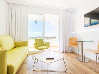 Espace salon lumineux dans une chambre d'hôtel avec canapé jaune, chaise, table et balcon avec vue sur la mer.