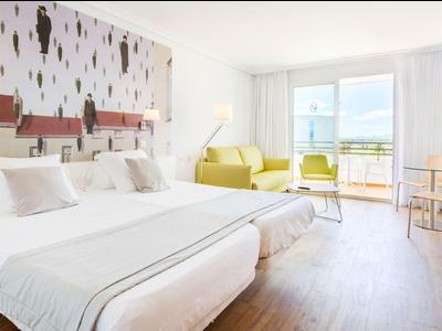 Chambre d'hôtel lumineuse avec deux lits simples, canapé jaune et balcon avec vue sur la mer.