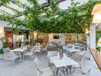 Terrasse d'hôtel moderne et lumineuse avec tables blanches, chaises et plantes vertes au plafond.
