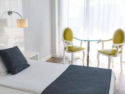 Chambre d'hôtel lumineuse avec lit simple, couverture grise et deux chaises vertes près de la fenêtre.