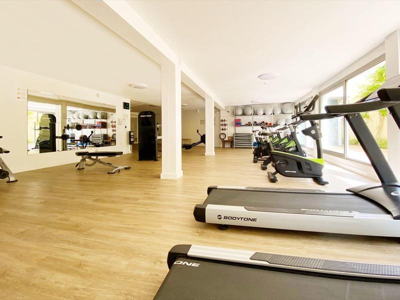 Salle de sport lumineuse avec tapis de course, haltères et grandes fenêtres laissant entrer la lumière.