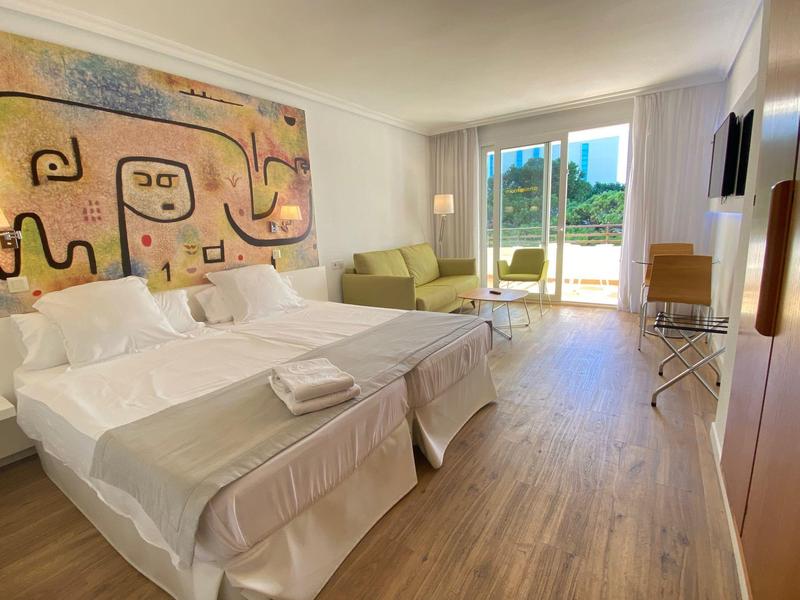 Chambre d'hôtel lumineuse avec deux lits, canapé, œuvre murale moderne et balcon avec vue sur la ville.