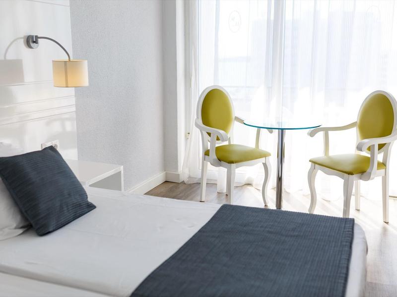 Chambre d'hôtel lumineuse avec lit simple, couverture grise et deux chaises vertes près de la fenêtre.