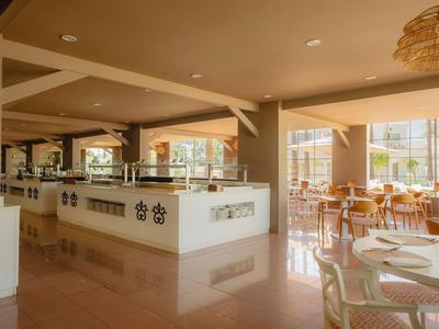 Helles Restaurant mit Sandtönen, großen Fenstern und Holzstühlen, heller Theke und Tischdekoration.