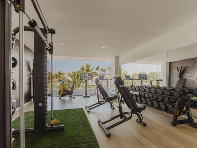Heller Fitnessraum mit Hantelbank, Gewichten, Kabelzug und großen Fenstern mit Außenblick.