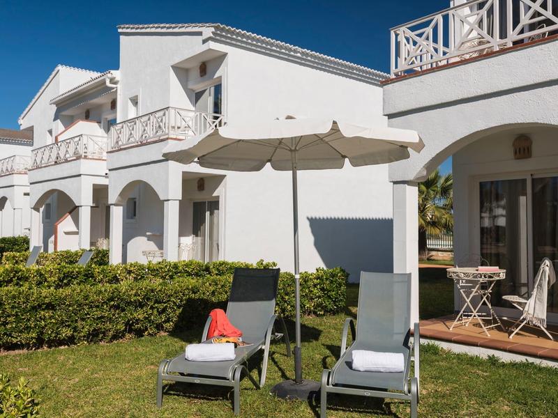 Bílý rekreační dům s terasou, slunečníkem a dvěma lehátky na zahradě.