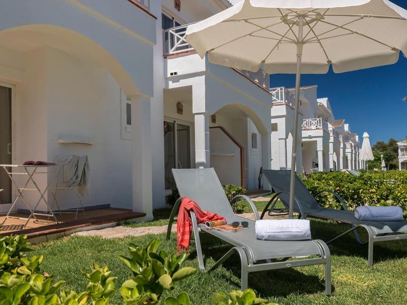 Bílé lehátka s deštníky před bílou hotelovou budovou na slunné zahradě.