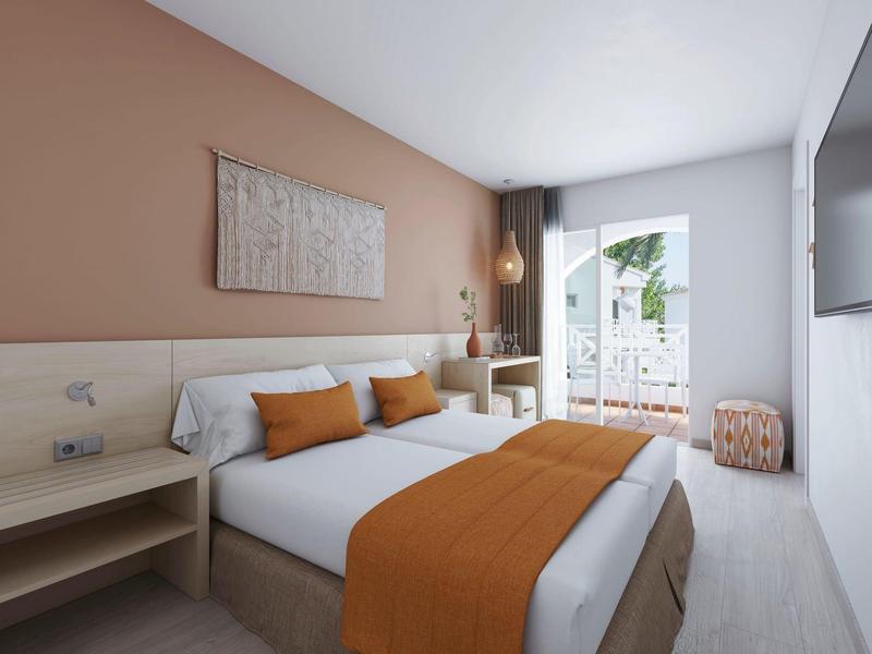 Světle hotelový pokoj s manželskou postelí, oranžovými akcenty a balkonem s rostlinami.