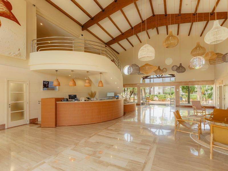 Světlá hotelová hala s recepcí, zaoblenou galerií a moderním osvětlením.