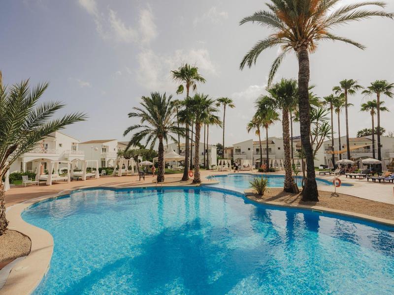 Hotelový bazén s palmami a lehátky pod modrou oblohou.