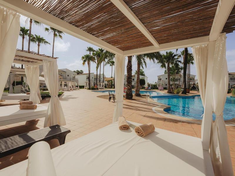 Pohled z krytého lehátka u bazénu na bazén a palmy v teplé atmosféře.