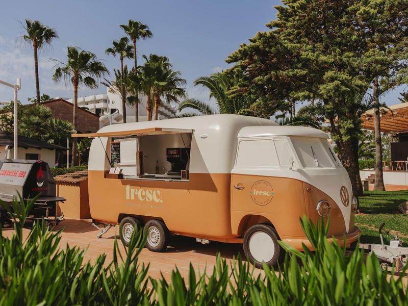 Malý retro oranžovo-bílý karavan před palmami a budovami.