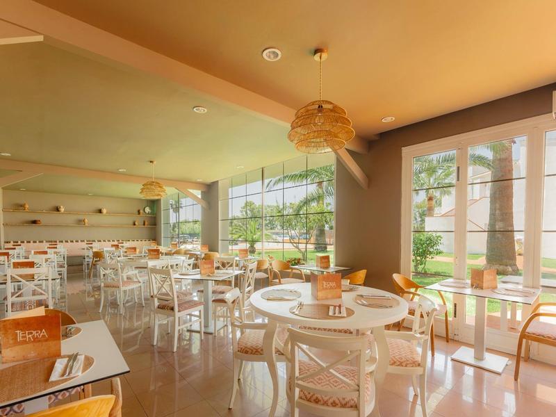 Světelná restaurace s bílými židlemi, kulatými stoly a velkými okny s výhledem na zeleň.