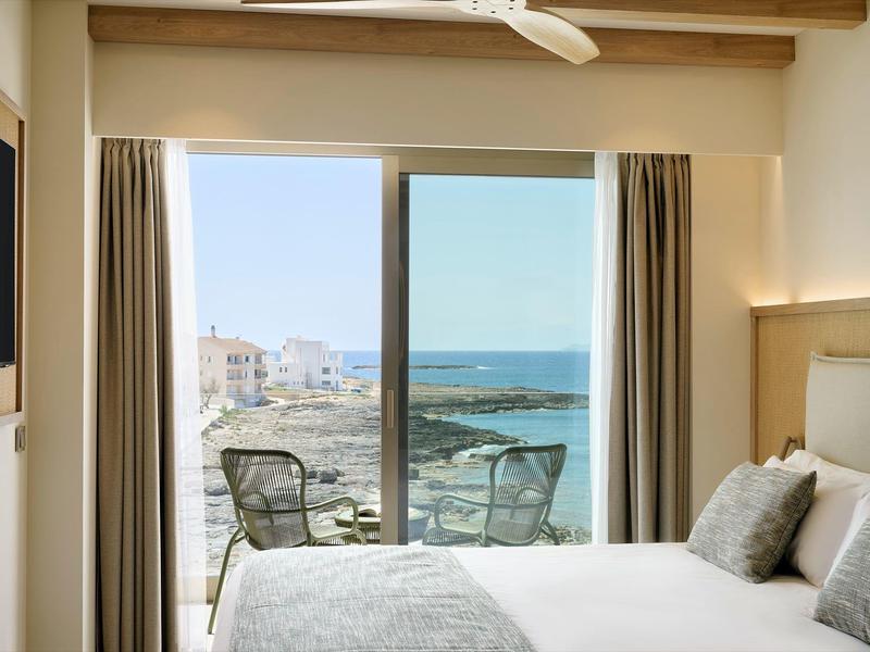 Helles Schlafzimmer mit weißer Bettwäsche und Balkon mit Stühlen und Meerblick im Hintergrund.