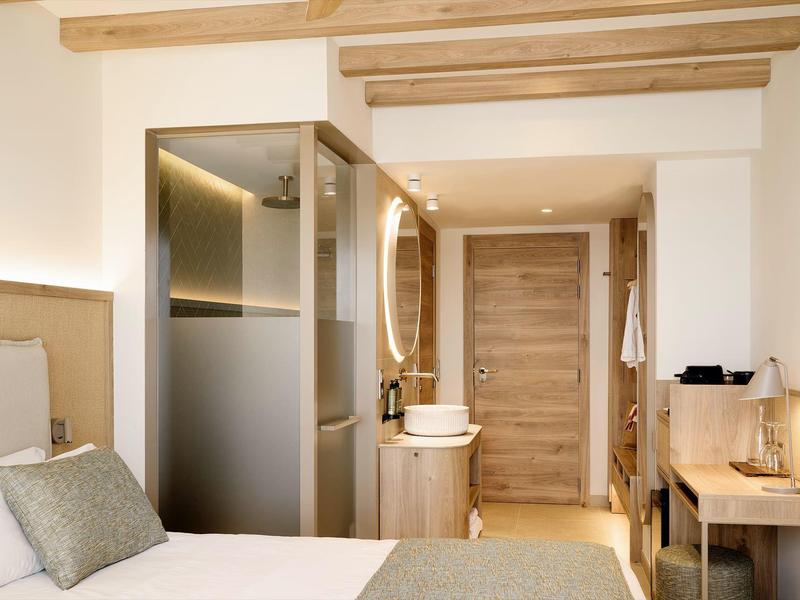 Helles Hotelzimmer mit weißem Bett, Holzboden, Glasschrank, Waschbecken und Schreibtisch aus Holz.