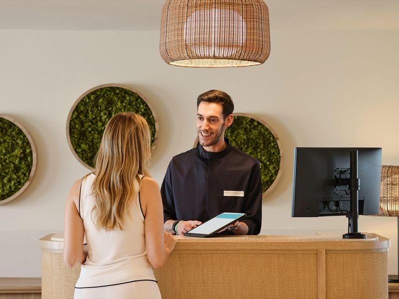 Ein Mann am Empfangstresen spricht mit einer Frau, moderner Raum mit Moosbildern an der Wand.