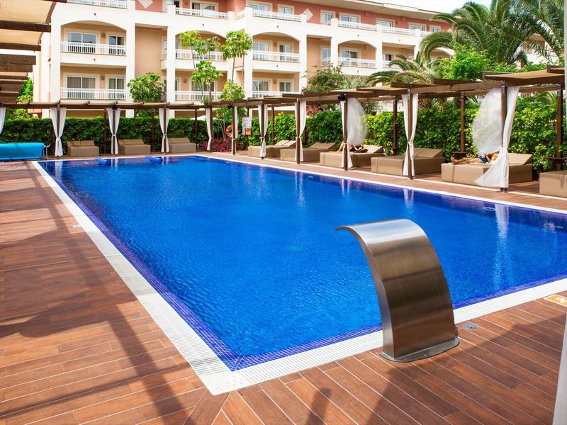 Moderner Außenpool mit klarem Wasser und Holzdeck vor einem Hotel mit Pflanzen.