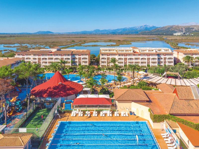 Großes Hotelresort mit mehreren Pools, roten Sonnensegeln und Blick auf Berge und Wasser
