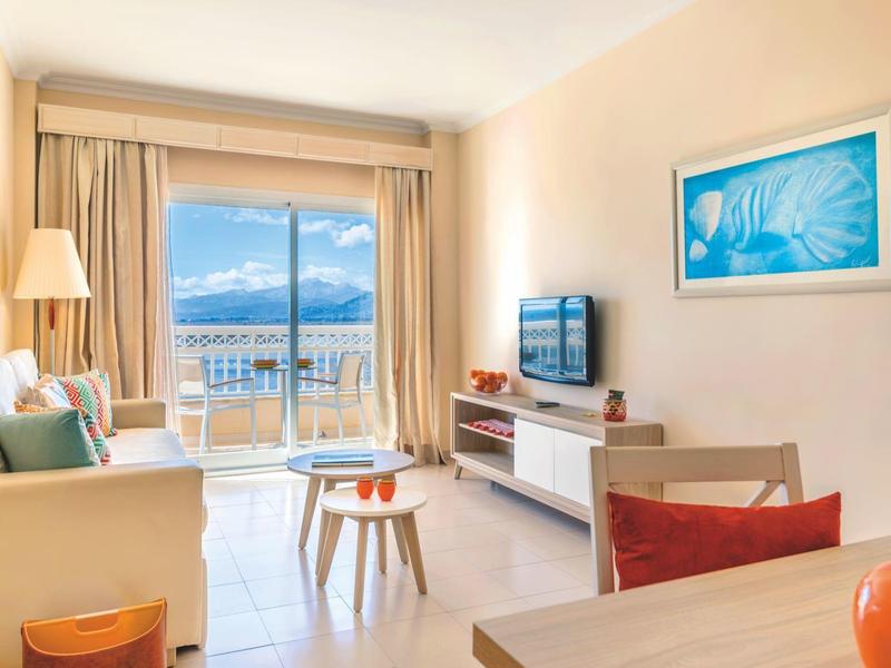 Helles Wohnzimmer mit Sofa, TV, Tisch und Blick auf Meer durch große Fenster.