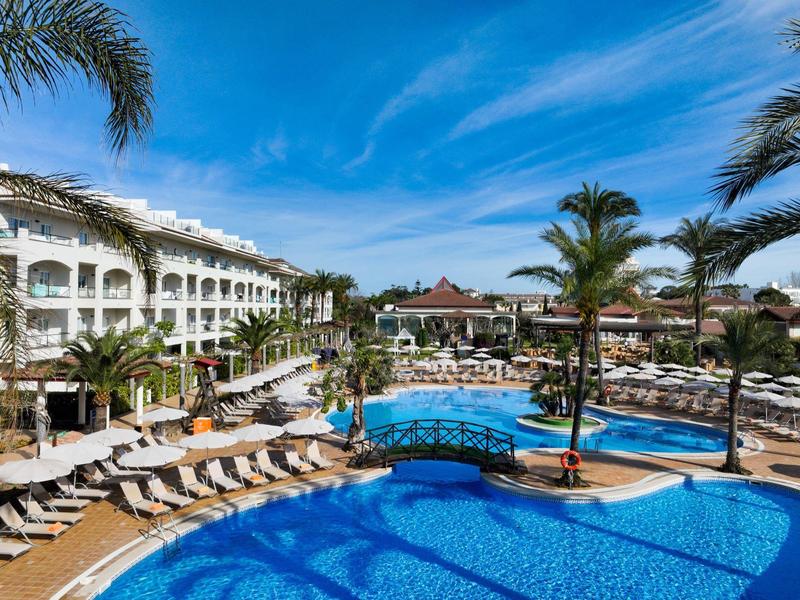 Großer Hotelpool mit Liegestühlen, Palmen und direktem Meerblick unter blauem Himmel.