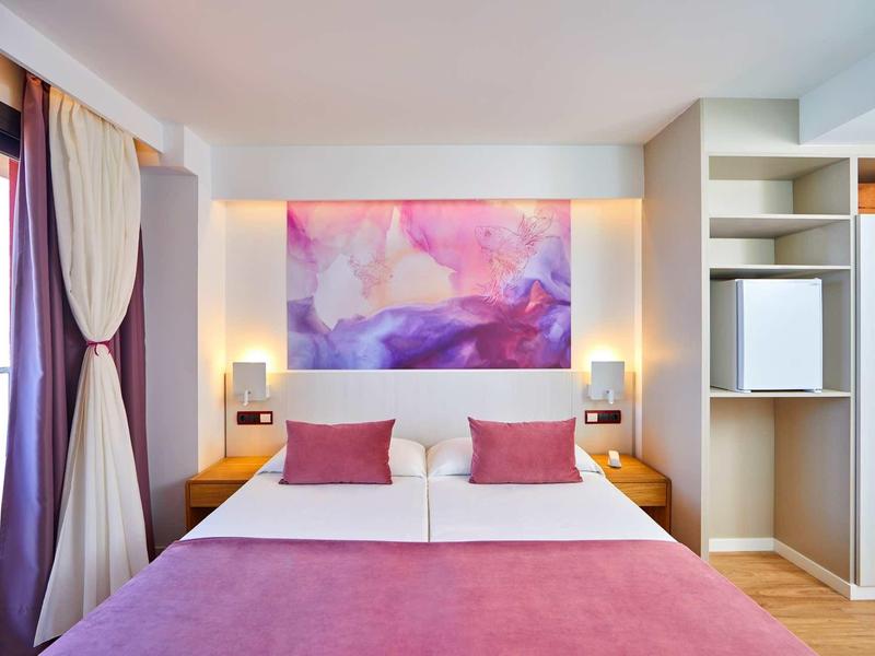 Modernes Schlafzimmer mit großem Bett, rosa Bettdecke, Kissen und abstraktem Bild über dem Kopfteil.