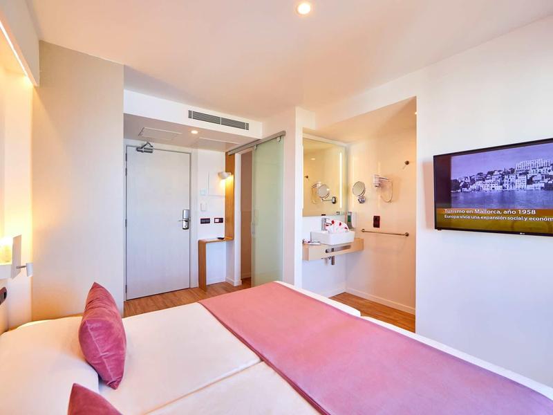 Helles Hotelzimmer mit Doppelbett, rosa Decke, großer TV und offenem Badezimmerbereich.