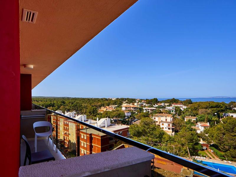 Balkon mit Glasgeländer, weißem Stuhl, roter Wand und Blick auf grüne Bäume und blauen Himmel.